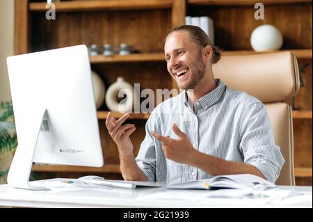Successful handsome confident young adult caucasian business man, manager or ceo with beard sitting in office, using computer, video communication with colleagues or clients, gesturing with hands Stock Photo