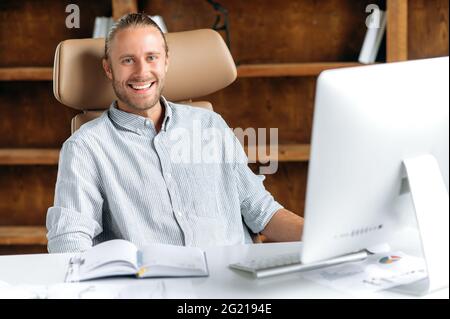 Portrait of happy successful smart young adult Caucasian manager, businessman or CEO with beard, wearing stylish shirt, sitting in office, using computer for work, looking at camera, smiling Stock Photo
