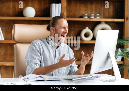 Frustrated angry caucasian businessman or CEO with a beard sits in the office, using a computer, yelling at employees via video link, crashed, loss or thwarted financial deal, gesturing with his hands Stock Photo