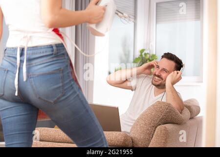 Young man sitting in living room taking off headphones smiling laughing. Woman standing holding mixer doing housework. Stock Photo
