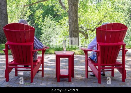 Rear view of older couple - man and woman - in their 70's, sitting in red Muskoka or Adirondack chairs on backyard patio overlooking trees in summer. Stock Photo