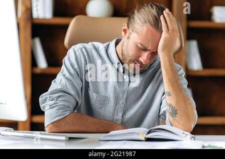 Tired overworked caucasian businessman, manager, office employee, sits at table in office, taking break from work, leaning his head on hand and closing his eyes, experiencing headache, migraine Stock Photo