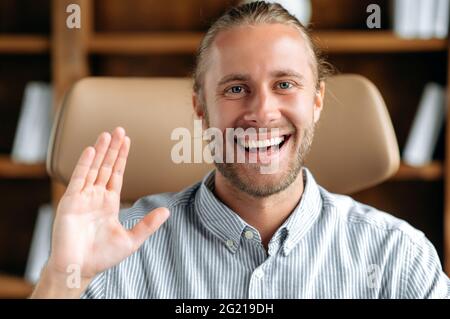 Portrait friendly successful smart confident caucasian manager, businessman or ceo, communicates with colleagues or client via video link, looks at camera, greets interlocutor with hand gesture, smile Stock Photo