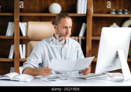 Smart focused young adult caucasian man, sits at the work desk in office, in stylish clothes, uses a computer, looks through documents and charts, thinks over financial strategy, working concentrated Stock Photo