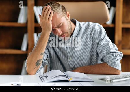 Young overwhelmed depressed caucasian businessman, manager or CEO, sitting at table in office, resting from work, propping up his head, closing his eyes, suffering from headache, migraine, needs rest Stock Photo