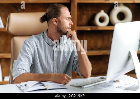 Pensive focused intelligent young adult caucasian businessman, manager or ceo, in stylish clothes, sitting at a table in the office, looking to the side, thinks over a business strategy and plans Stock Photo
