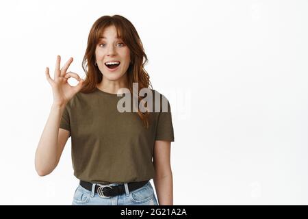 Very good, awesome. Smiling happy girl showing OK okay sign, say yes and looking amazed, praise choice, making compliment, positive feedback, standing Stock Photo