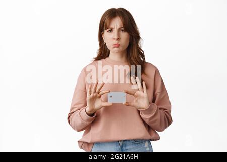Portrait of young woman bank client, looks disappointed and pouting, shows credit card upset, stands against white background Stock Photo