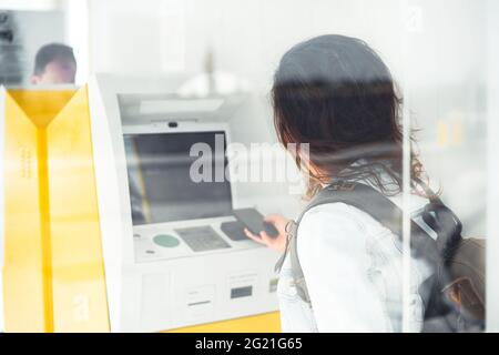Pretty young woman traveler wearing protective face mask paying with phone at yellow ATM. Modern banking technology helping tourists in easy payments Stock Photo