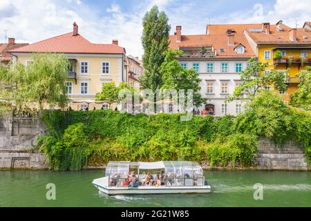 Ljubljana, Slovenia - August 15, 2018: A tourist boat passes on Ljubljanica river beside old picturesque buildings and green vegetation in downtown Stock Photo