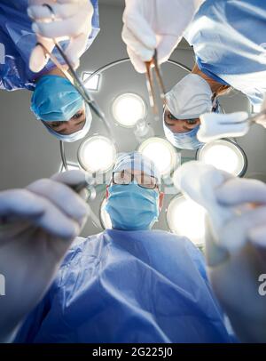 View from below of doctors holding scalpel, scissors and forceps with tampon while looking at patient during medical operation. Team doing plastic surgery in operating room. Concept of medicine Stock Photo