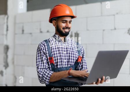 Portrait of development supervisor wearing protective vest and helmet over formal suit proofing blueprints using laptop computer inside unfinished bui Stock Photo