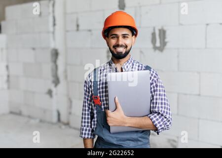 Portrait of development supervisor wearing protective vest and helmet over formal suit proofing blueprints using laptop computer inside unfinished bui Stock Photo