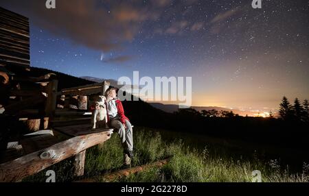 Woman traveler hugging cute dog while enjoying fantastic view of night starry sky. Female hiker with four-legged friend sitting on wooden stairs under majestic sky with stars and comet Neowise. Stock Photo