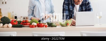 Couple enjoying and preparing healthy meal in kitchen and reading recipes on laptop Stock Photo