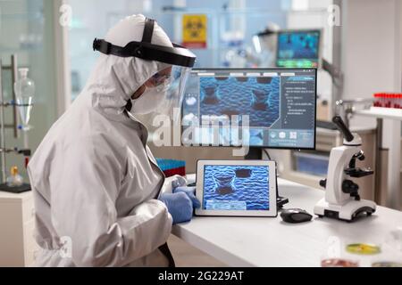 Laboratory chemist wearing ppe suit using tablet pc with virus on display. Scientist examining virus evolution using high tech technology researching vaccine development against covid19. Stock Photo