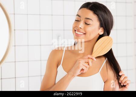 smiling young african american woman brushing hair with closed eyes near mirror in bathroom Stock Photo