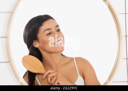 smiling young african american woman brushing hair near mirror in bathroom Stock Photo