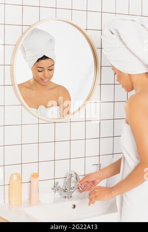 young african american woman wrapped in towels standing near washbasin and touching metal faucet in bathroom Stock Photo