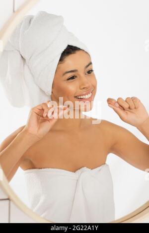 smiling young african american woman wrapped in towels holding dental floss near face and looking in mirror in bathroom Stock Photo
