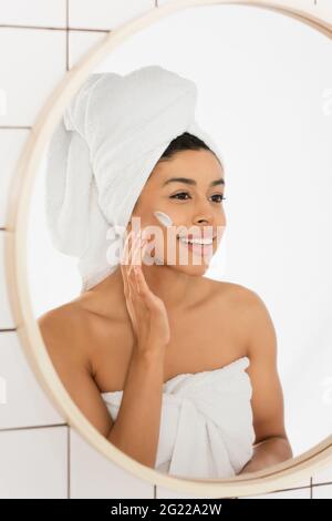 young african american woman wrapped in towels applying cream on face and looking in mirror in bathroom Stock Photo