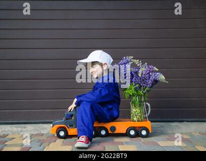 funny cute 4-5 year old boy in a blue mechanic overalls and a cap sits in profile on a toy truck with a large bouquet of flowers. Delivery concept, li Stock Photo