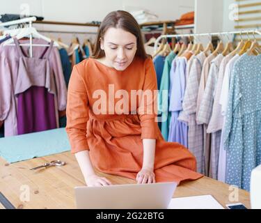 Working on new collection. Young successful business woman fashion designer sitting on table and works on laptop at her own workshop, female dressmaker or seamstress creating clothing design online Stock Photo