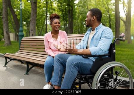 Happy handicapped black guy in wheelchair talking to his girlfriend at park, copy space Stock Photo