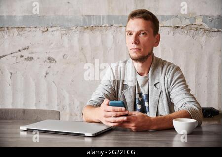 Portrait of handsome young man student with beard sitting with laptop and cup coffee and holding smartphone. Concept of working or studying in phone. Stock Photo