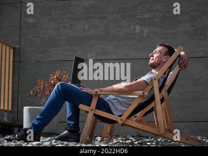 Side view of smiling young man sitting on folding chair and enjoying sunny weather on street. Concept of process working or studying in laptop at home outdoors. Stock Photo