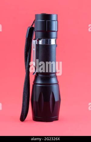 A black flashlight of powerful light standing on a red background. The tactical flashlight is off, has a tether and a hook so that it can be hung from Stock Photo