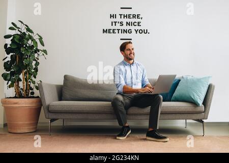 Confident executive sitting on sofa using laptop at office lobby. Happy man working on laptop and looking away. Stock Photo