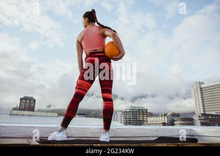Rear view of woman in sportswear exercising with a basketball with  cityscape in background. fitness woman holding a basketball standing on rooftop. Stock Photo