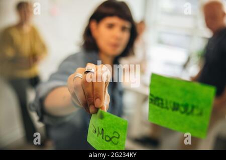 Close up shot of hand of woman sticking adhesive notes on glass wall in office. Female professional presenting ideas to colleagues during meeting. Stock Photo