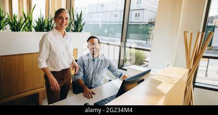 Mature woman standing and young man sitting at desk smiling at camera. business executives looking at camera. Stock Photo