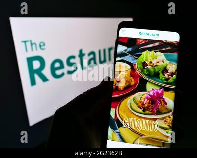 Person holding cellphone with webpage of British gastronomy company The Restaurant Group plc on screen with logo. Focus on center of phone display. Stock Photo