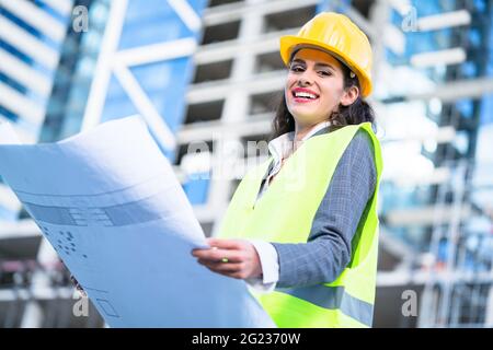 Female architect studying drafts while visiting large construction site Stock Photo