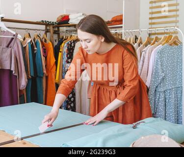 Focused young woman seamstress marking material with piece of soap for sewing clothes, dressmaker working on new dress collection in own atelier. Female owner of local clothing business at work Stock Photo