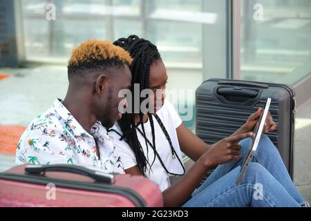 Young african couple checking their travel schedule on a tablet, sitting at the station with their suitcases. Vacation concept. Stock Photo