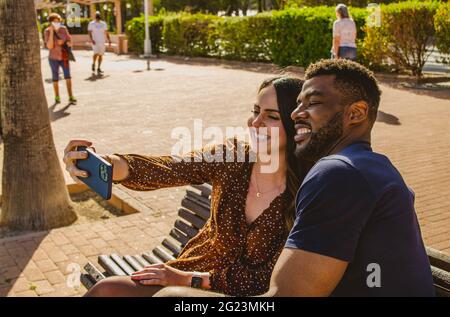Happy Multiethnic Couple Taking a Selfie with Phone Together Sitting on a Bench on the Promenade Dur Stock Photo