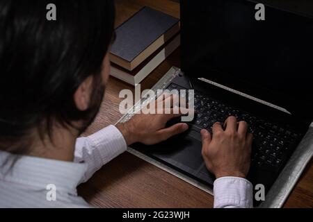 Hands of Long Haired Brown Hispanic Man Dressed in Shirt Typing on a Black Laptop on a Wooden Table Stock Photo