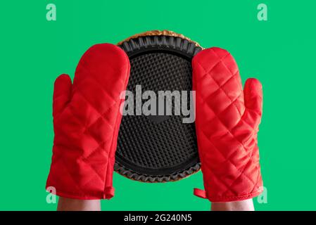 Woman hands protected with gloves taking out of oven baking tray. Holding baking tray with red gloves bottom-up view isolated on a green background. Stock Photo