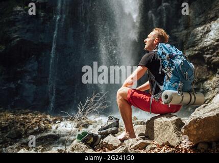 Man with backpack dressed in active trekking clothes takes off trekking boots sitting near mountain river waterfall and enjoying the Nature. Traveling Stock Photo
