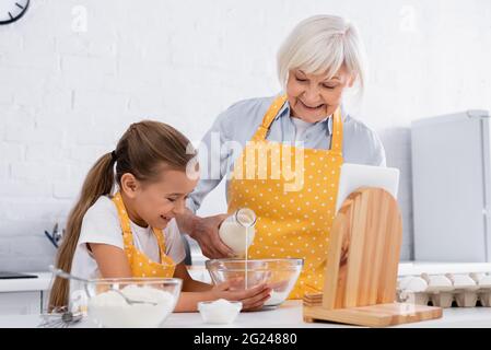 Positive kid and grandmother pouring milk in bowl near digital tablet and flour Stock Photo