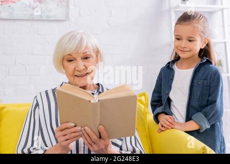 Smiling kid standing near grandmother reading book on couch Stock Photo