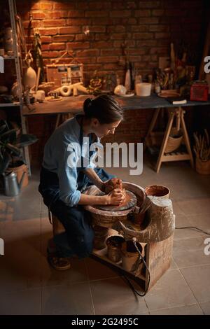 Happy cheerful young female artisan wearing apron shaping clay pot in pottery workshop Stock Photo