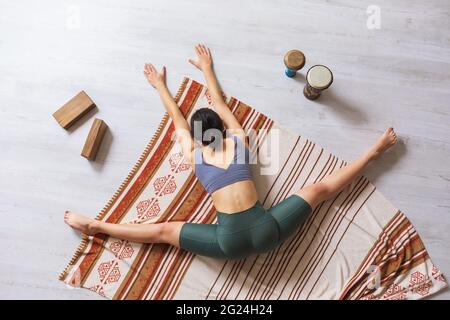 A woman in sportswear, doing yoga, performs a samokanasana exercise while sitting on a blanket, a transverse split with the body tilted forward. Stock Photo