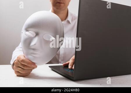 A man working at a laptop holds a mask in his hand, a concept on the topic of anonymity on the Internet. Stock Photo