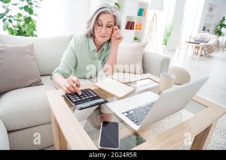 Photo of focused minded person sit on couch hand touch glasses look calculate results working from home indoors Stock Photo