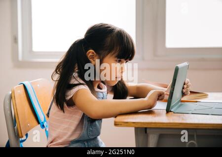 Asian school girl using digital device in school classroom, digital native, technology, learning, touchscreen. Female elementary student with tablet in class. Stock Photo